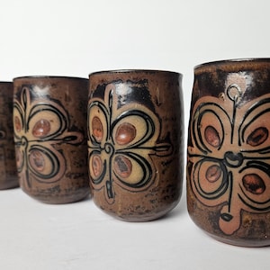 Puede incluir: Cuatro vasos de cerámica marrón con un diseño floral en tonos beige, negro y rojo. Los vasos tienen un aspecto rústico y hecho a mano y están dispuestos en fila. El diseño es simétrico y se repite en cada vaso.