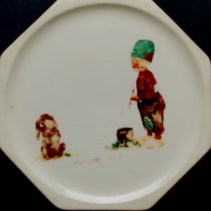 Puede incluir: Plato de cerámica blanco con una ilustración de un niño con un sombrero verde sosteniendo un palo y un perro sentado en el suelo. La ilustración está en tonos de verde, rojo y marrón.