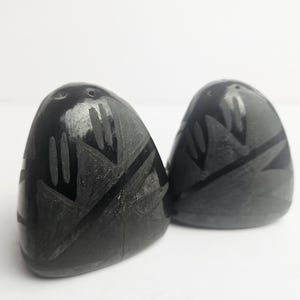 May include: Two dark gray, egg-shaped salt and pepper shakers. Each shaker has a carved geometric design in a darker shade of gray. The shakers are on a white surface.