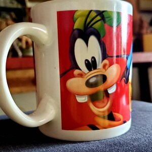 May include: White ceramic mug with a colorful cartoon image of Goofy on the front. Goofy is wearing a red shirt and has a big smile. The mug has a handle on the left side.