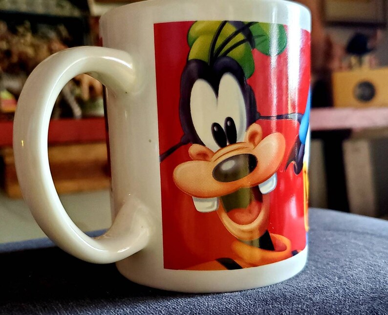 May include: White ceramic mug with a colorful cartoon image of Goofy on the front. Goofy is wearing a red shirt and has a big smile. The mug has a handle on the left side.