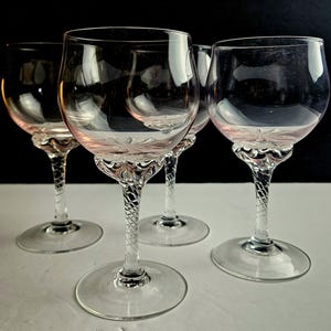 May include: Four clear wine glasses with a decorative floral design on the bowl and a twisted stem. The glasses are arranged on a white surface against a dark background. The glasses are empty.