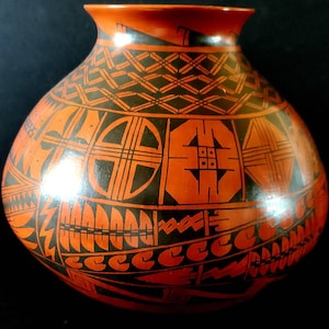 Op de afbeelding: Een handbeschilderde, ronde keramische vaas met een brede opening. De vaas heeft zwarte geometrische patronen op een oranje achtergrond, waaronder ruiten, kruisen en hoekige vormen. Het ontwerp bedekt het gehele oppervlak van de vaas.
