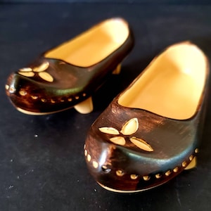 May include: A pair of miniature wooden shoes, each with a dark brown exterior and a light tan interior. The shoes have a floral design on the toe and are accented with small, light-colored dots. The heels are also light tan.