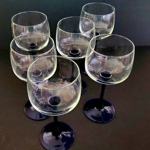 May include: Six clear glass wine glasses with blue stems. The glasses are arranged in a circle on a dark surface.