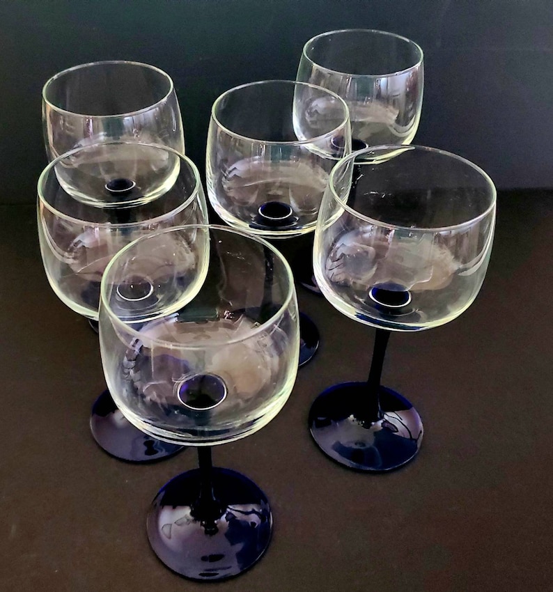 May include: Six clear glass wine glasses with blue stems. The glasses are arranged in a circle on a dark surface.