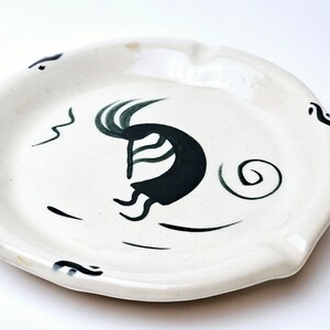 May include: White ceramic ashtray featuring a stylised black and green bird design in flight. The design includes a swirl and wavy lines.