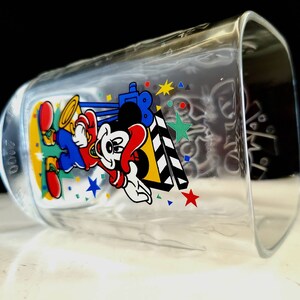 May include: A clear glass cup with a Mickey Mouse design. Mickey is wearing a red shirt, blue pants, and a yellow hat. He is holding a clapperboard and there are stars and confetti around him. The year 2000 is printed on the side of the cup.