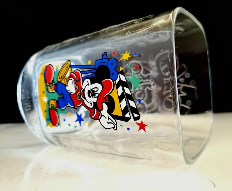 May include: A clear glass cup with a Mickey Mouse design. Mickey is wearing a red shirt, blue pants, and a yellow hat. He is holding a clapperboard and there are stars and confetti around him. The year 2000 is printed on the side of the cup.