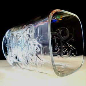 May include: Clear glass drinking glass with an etched design of stars and a stylized image of a person riding a horse. The glass has a square shape.