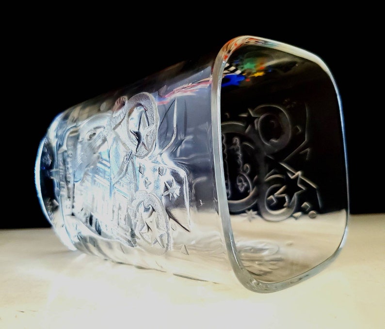 May include: Clear glass drinking glass with an etched design of stars and a stylized image of a person riding a horse. The glass has a square shape.