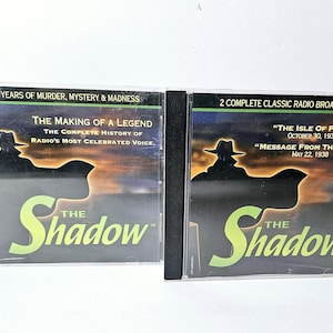 Puede incluir: Dos estuches de CD con The Shadow, un programa de radio clásico. El estuche de la izquierda presenta el texto "The Making of a Legend" y "The Complete History of Radio's Most Celebrated Voice." El estuche de la derecha presenta el texto "The Isle of Fear" y "Message From the Hills."