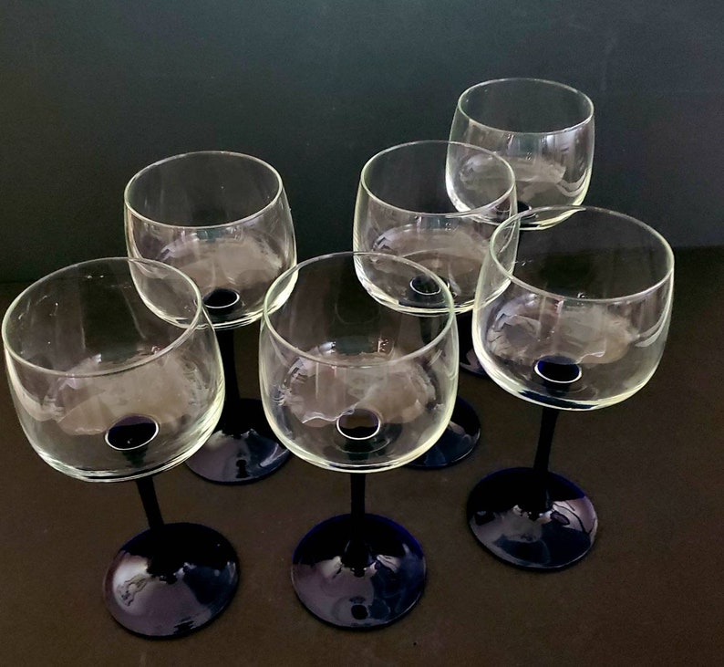 May include: Six clear glass wine glasses with blue stems. The glasses are arranged in a circle on a dark surface.