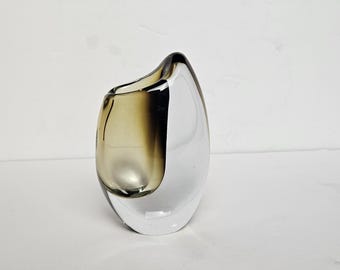 May include: A modern art glass vase with a unique, asymmetrical shape. The vase is a combination of clear and smoky brown glass, creating a two-tone effect. The design features a curved opening and a rounded base, suitable for home decor.