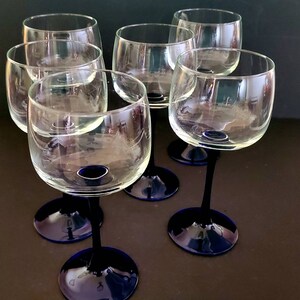 May include: Six clear glass wine glasses with blue stems and bases. The glasses are arranged in a semi-circle on a dark surface.