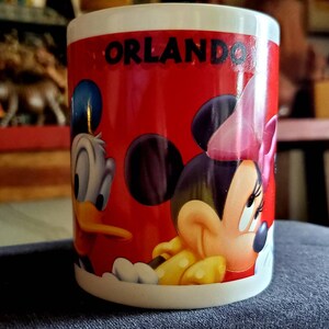 May include: A white ceramic mug with a red background and a picture of Mickey Mouse and Donald Duck. The mug says "Orlando" in black letters.