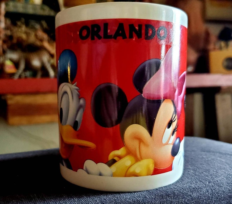 May include: A white ceramic mug with a red background and a picture of Mickey Mouse and Donald Duck. The mug says "Orlando" in black letters.