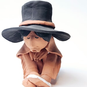 Puede incluir: Una figurita de terracota de una persona que lleva un sombrero negro y un abrigo marrón, sentada con las piernas cruzadas y la cabeza gacha.