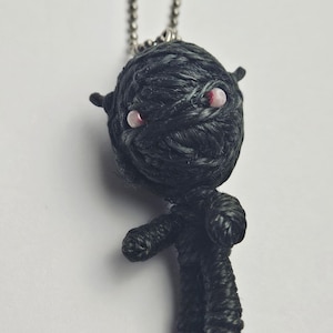 May include: A handmade black yarn doll with red bead eyes and a silver chain. The doll has a wrapped head, small arms, and legs. The doll is a keychain and is set against a white background.