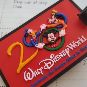 Może przedstawiać: Czerwona etykieta bagażowa z Myszka Miki, Kaczorem Donaldem i Goofym. Etykieta zawiera napisy "Walt Disney World" w kolorze białym i niebieskim, z tekstem "Celebrate the Future Hand in Ha". Numer "2" jest żółty.