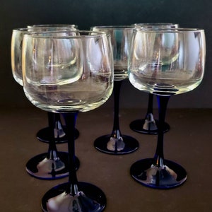 May include: Six clear glass wine glasses with blue stems. The glasses are arranged in a semi-circle on a brown surface.