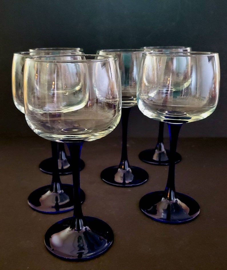 May include: Six clear glass wine glasses with blue stems. The glasses are arranged in a semi-circle on a brown surface.