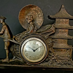 Op de afbeelding: Vintage klok in bronskleur met een Japanse scène. De wijzerplaat is omgeven door een gedetailleerd reliëf van een riksja met een passagier, een riksjatrekker en een pagode. De wijzerplaat heeft een zilveren afwerking met zwarte cijfers.
