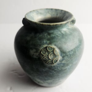 Op de afbeelding: Een bolvormige keramische vaas met een gevlekt glazuur in blauwgroen en donkerblauw. De vaas heeft een brede, uitlopende rand en een klein, rond medaillon aan de voorzijde dat is versierd met een verhoogd, herhalend spiraalpatroon in lichtblauw.