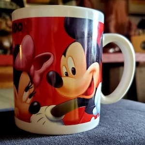 May include: A white ceramic mug with a red background and a cartoon image of Mickey Mouse and Minnie Mouse. Mickey Mouse is on the right side of the mug and Minnie Mouse is on the left side. Mickey Mouse is wearing red shorts and Minnie Mouse is wearing a red dress with a pink bow.