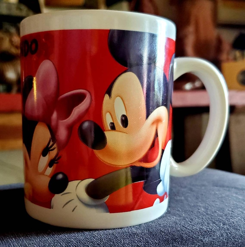 May include: A white ceramic mug with a red background and a cartoon image of Mickey Mouse and Minnie Mouse. Mickey Mouse is on the right side of the mug and Minnie Mouse is on the left side. Mickey Mouse is wearing red shorts and Minnie Mouse is wearing a red dress with a pink bow.