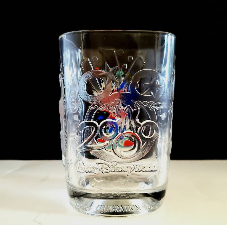 May include: Clear glass tumbler with a frosted design featuring the Walt Disney World logo and the year 2000. The design includes a Mickey Mouse silhouette and other celebratory imagery.