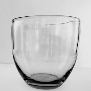 Könnte beinhalten: Ein klares, abgerundetes Glas mit einem leicht unregelmäßigen Rand. Das Glas hat eine weite Öffnung und einen geschwungenen, bauchigen Körper. Das Glas ist transparent und lässt Licht durch. Es wird wahrscheinlich zum Trinken verwendet und steht auf einer weißen Oberfläche.
