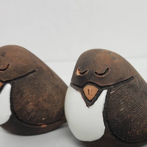 Könnte beinhalten: Zwei Keramik-Vogelfiguren mit geschlossenen Augen und weißem Bauch. Die Vögel sind braun mit einer strukturierten Oberfläche.