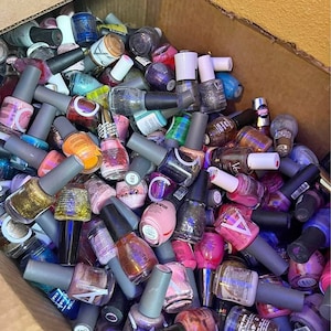 May include: A cardboard box overflowing with various nail polish bottles. The bottles have different colors, including pink, gold, blue, and purple. The bottles have gray or white caps. Some bottles have text on them.