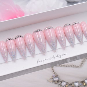 May include: A set of ten stiletto-shaped press-on nails featuring a light pink base with a silver holographic glitter gradient fading toward the tips. Each nail is accented with small silver rhinestones at the cuticle line. Text reads baroquenails.etsy.com.