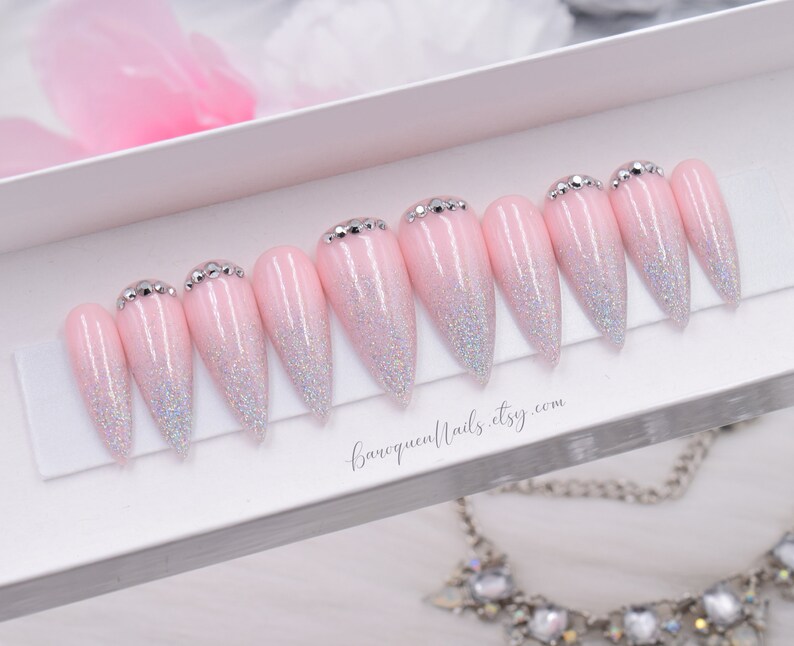 May include: A set of ten stiletto-shaped press-on nails featuring a light pink base with a silver holographic glitter gradient fading toward the tips. Each nail is accented with small silver rhinestones at the cuticle line. Text reads baroquenails.etsy.com.