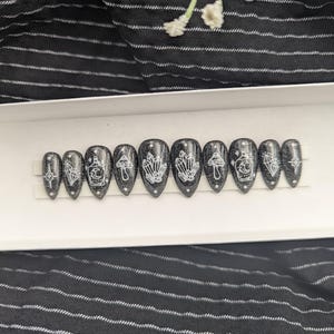 May include: A set of ten almond-shaped press-on nails with a black glitter finish. Each nail features white line art designs including crystals, mushrooms, potion bottles, and stars. The nails are arranged in a row on a white display strip.