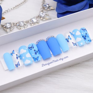 Puede incluir: Un conjunto de uñas postizas azules con diseños de mariposas y nubes. Algunas uñas son de color azul liso, mientras que otras presentan nubes blancas y mariposas azules. Una uña tiene una fila de pedrería.