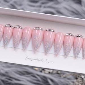 May include: A set of ten stiletto-shaped press-on nails in a light pink base color with a silver holographic glitter gradient starting from the tips. Each nail features a row of small clear rhinestones at the cuticle line. The text baroquennails.etsy.com is printed below the nails.