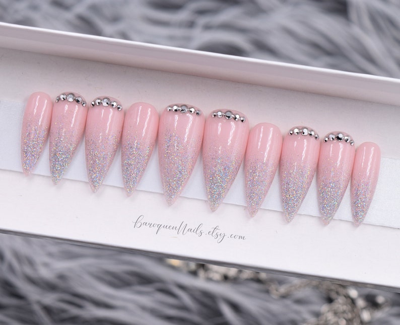 May include: A set of ten stiletto-shaped press-on nails in a light pink base color with a silver holographic glitter gradient starting from the tips. Each nail features a row of small clear rhinestones at the cuticle line. The text baroquennails.etsy.com is printed below the nails.