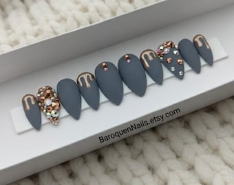 May include: A set of ten matte charcoal gray press-on nails in a stiletto shape, displayed in a white box with the text BaroquenNails.etsy.com. The nails feature metallic gold drip accents, clear crystals, and bronze-toned rhinestones.