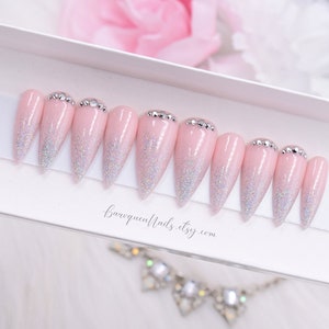 May include: A set of ten stiletto-shaped press-on nails in a light pink shade with a silver holographic glitter gradient starting from the tips. Each nail features a row of small clear rhinestones at the cuticle base. The text baroquenails.etsy.com is visible.