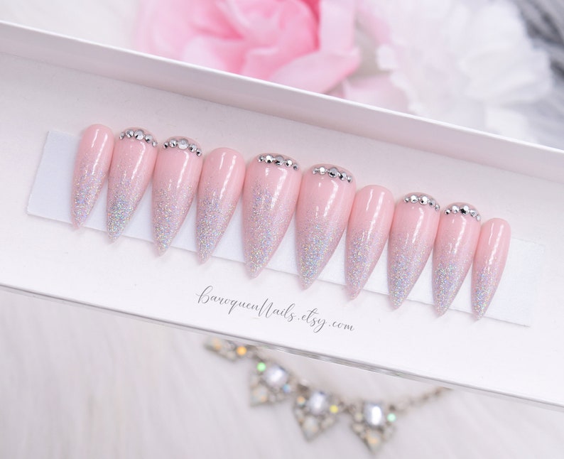 May include: A set of ten stiletto-shaped press-on nails in a light pink shade with a silver holographic glitter gradient starting from the tips. Each nail features a row of small clear rhinestones at the cuticle base. The text baroquenails.etsy.com is visible.