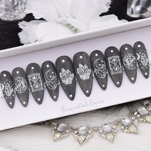 May include: A set of ten stiletto-shaped press-on nails in a dark charcoal gray glitter finish. Each nail features white line art designs including crystals, moons, stars, and an hourglass. The text BaroqueNails.etsy.com is printed on the white display card.