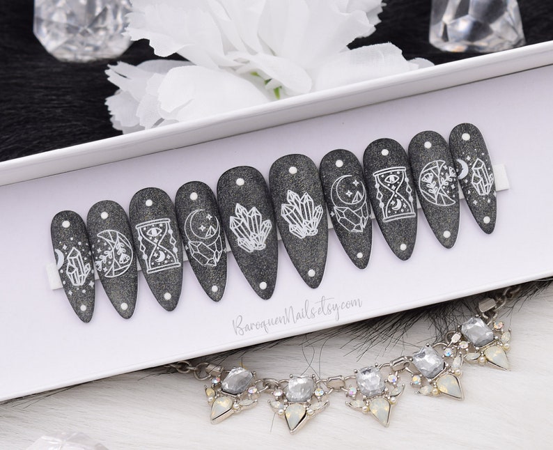 May include: A set of ten stiletto-shaped press-on nails in a dark charcoal gray glitter finish. Each nail features white line art designs including crystals, moons, stars, and an hourglass. The text BaroqueNails.etsy.com is printed on the white display card.