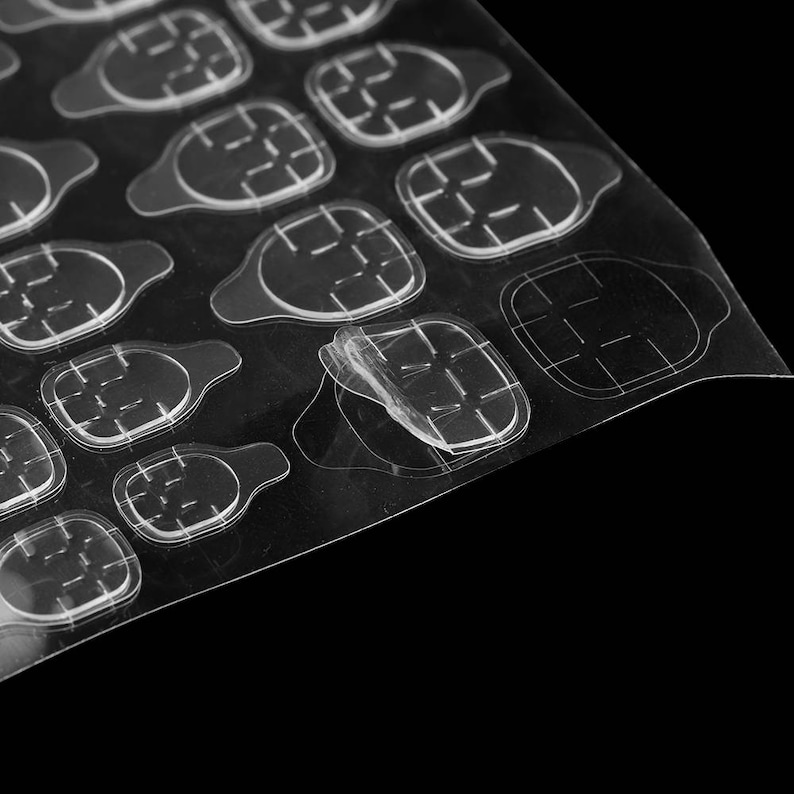 May include: A sheet of clear, double-sided adhesive nail tabs arranged in rows. Each individual tab features a rounded, shield-like shape with a small pull tab at the top, designed for attaching artificial nails to natural fingernails.