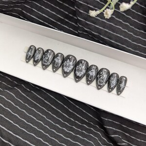 May include: A set of ten almond-shaped press-on nails featuring a black glitter base with white line art designs. The patterns include crystals, mushrooms, and potion bottles with celestial motifs, arranged in a row on a white display strip.