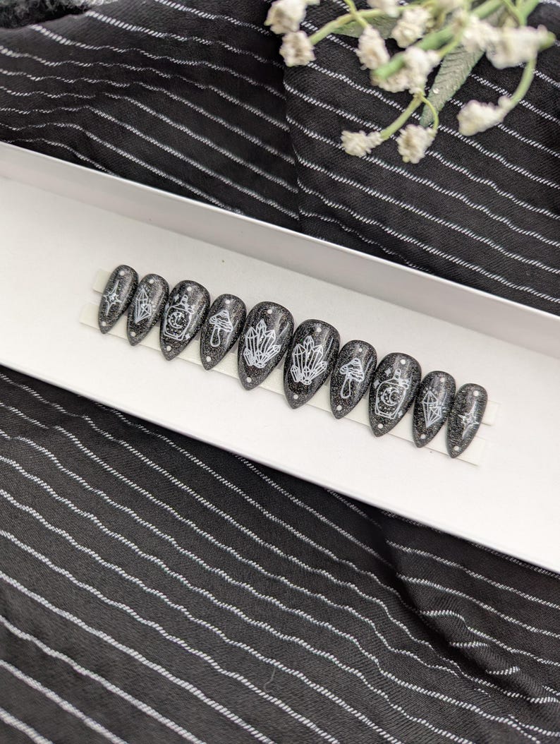 May include: A set of ten almond-shaped press-on nails featuring a black glitter base with white line art designs. The patterns include crystals, mushrooms, and potion bottles with celestial motifs, arranged in a row on a white display strip.