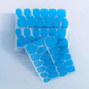May include: A collection of bright blue adhesive nail tabs arranged on clear plastic sheets. The set includes various sizes of pre-cut, flexible, double-sided sticky tabs designed for attaching artificial nails, presented in multiple rows.