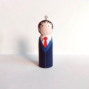 Könnte beinhalten: Ein handbemaltes Holz Ornament, das eine Person in einem blauen Anzug und einer roten Krawatte darstellt. Das Ornament hat eine Schlaufe oben zum Aufhängen.